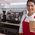 Happy young barista offering cup of coffee to go smiling at came
